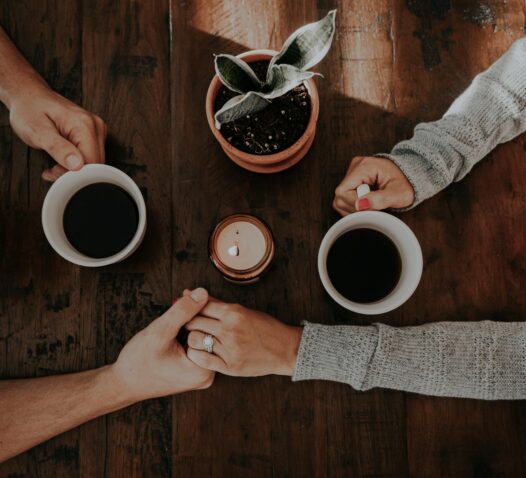 Två personer håller varandra i händerna över en träbord med två kaffekoppar och en liten växt.