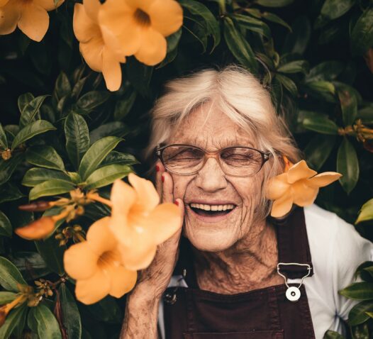 En leende äldre kvinna omgiven av orange blommor och gröna blad.